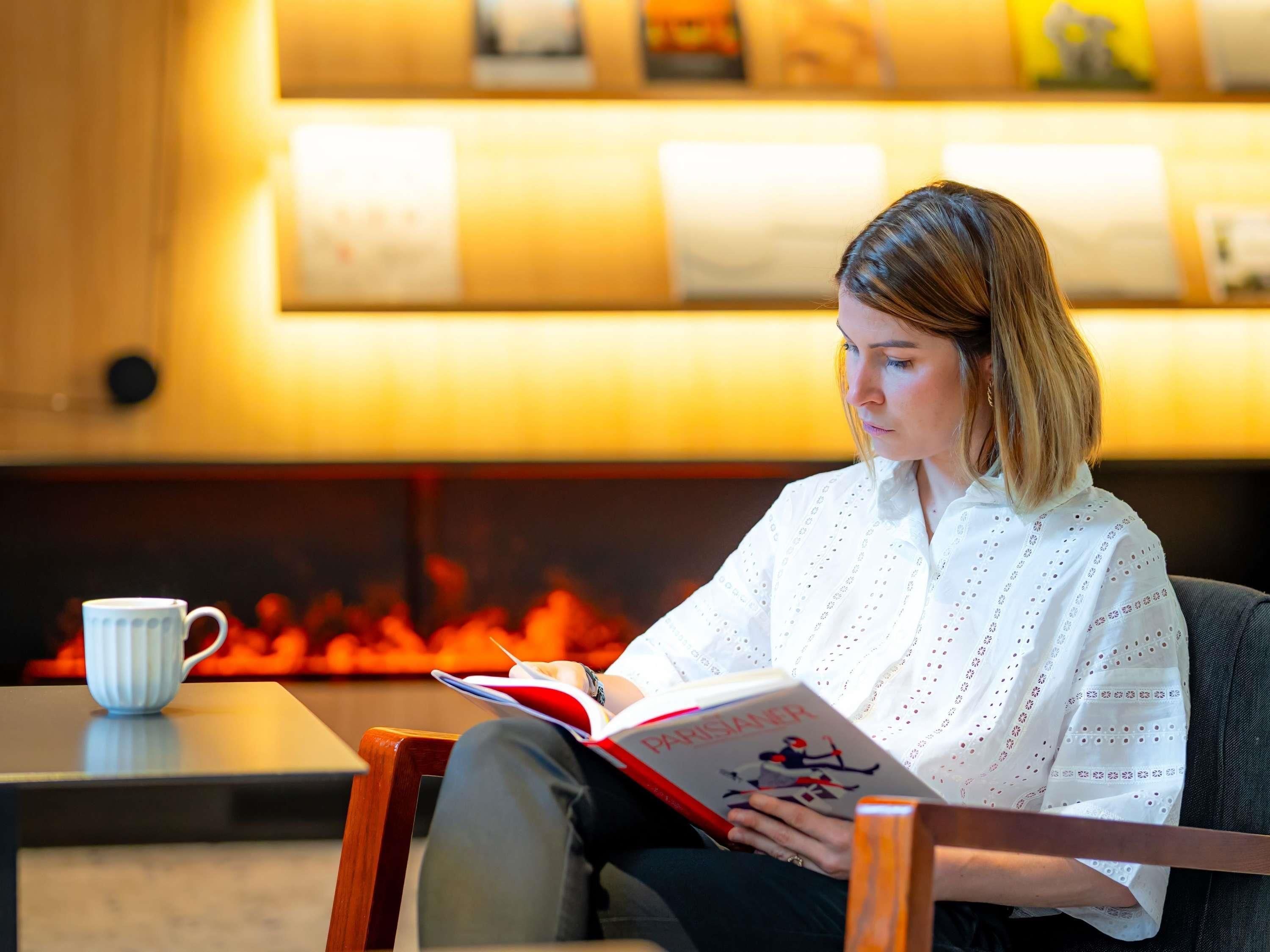 Une personne assise sur une chaise, lisant un livre, avec une tasse posée sur une table.