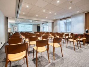 Un salón de conferencias con filas de sillas, una pantalla de proyección y un ventanal con cortinas.