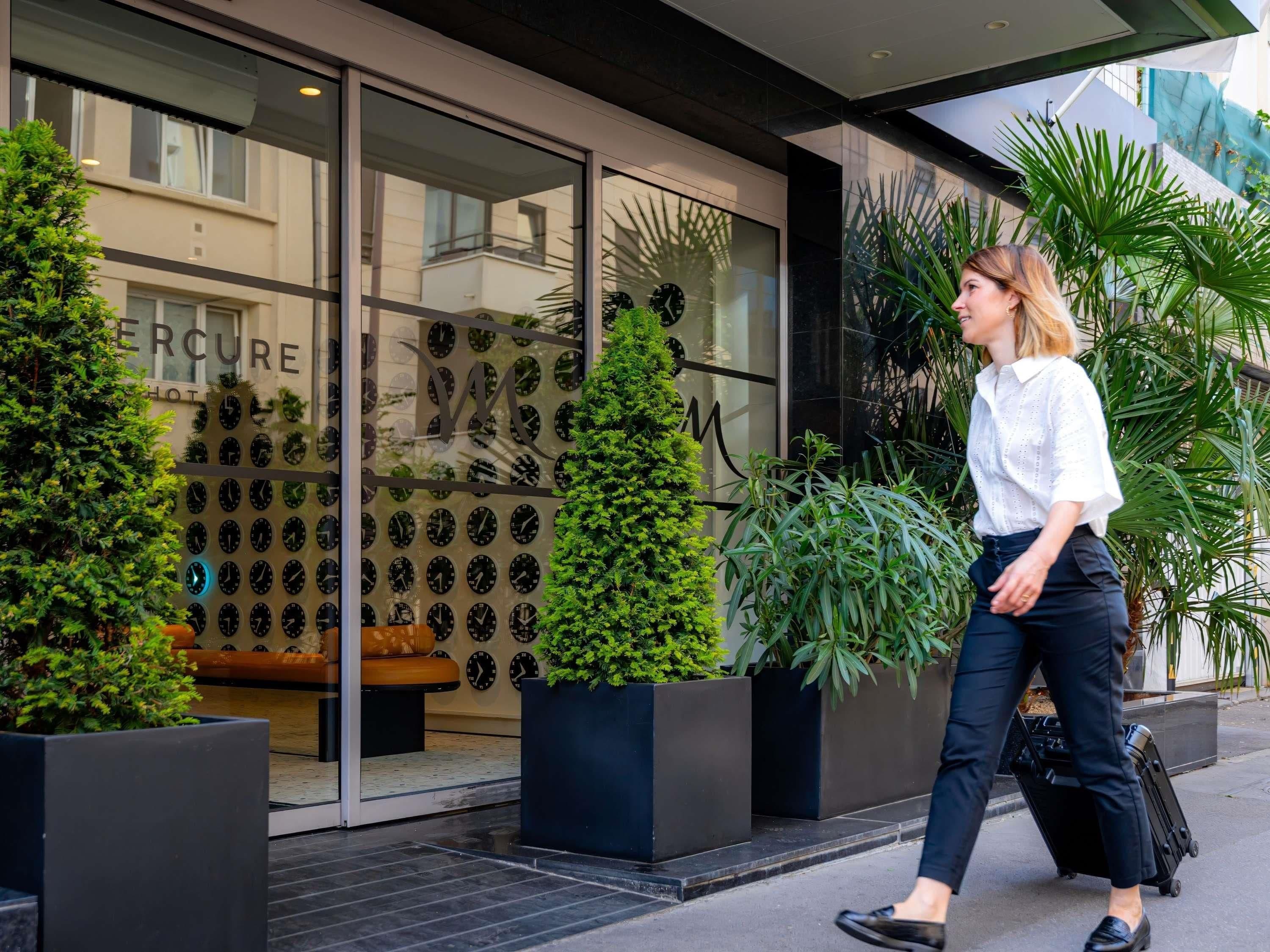 L’entrée d’un hôtel avec une façade en verre, un mur décoré d’une horloge et des plantes en pot.