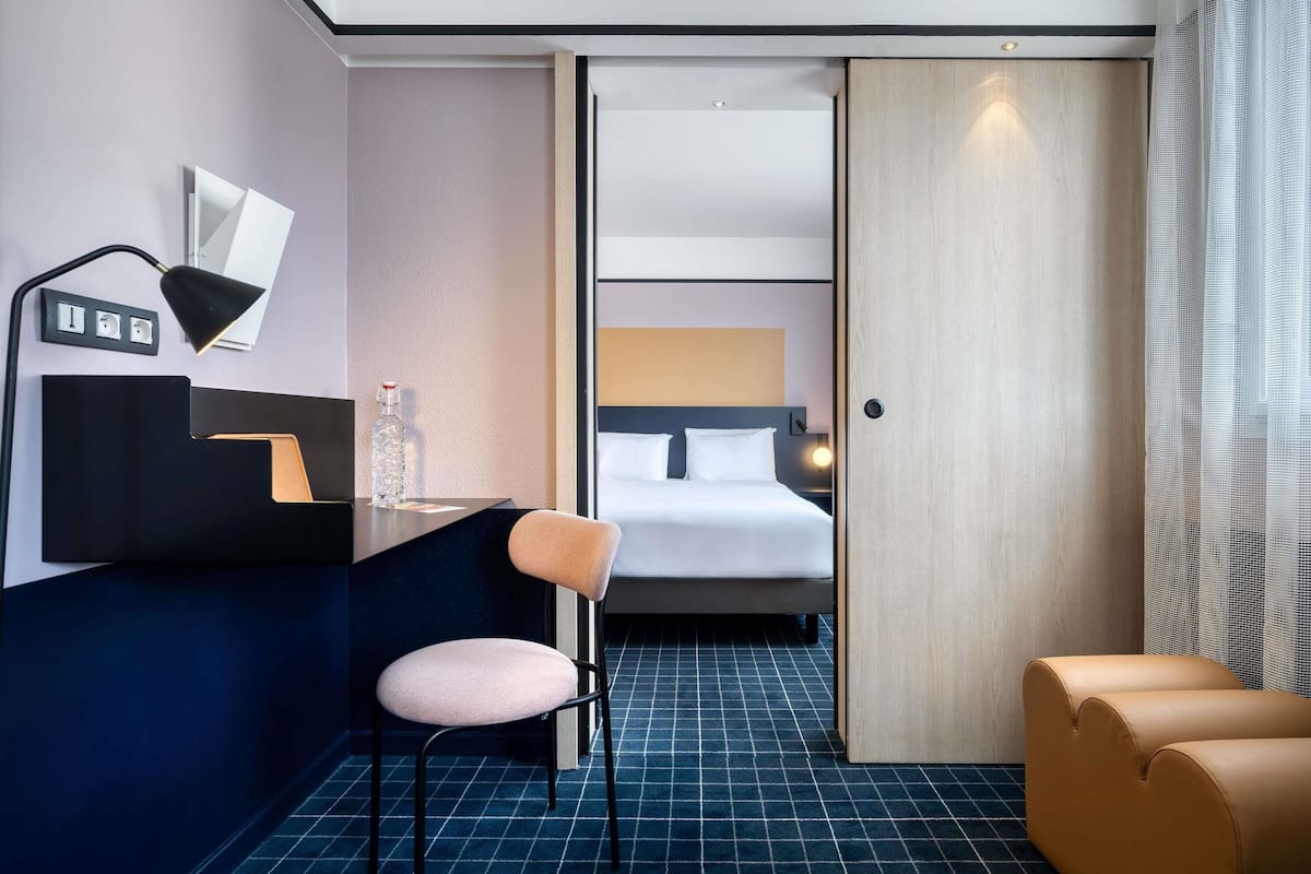 A hotel room with a bed, a desk with a chair, a small table with a bottle, and a wall-mounted lamp.