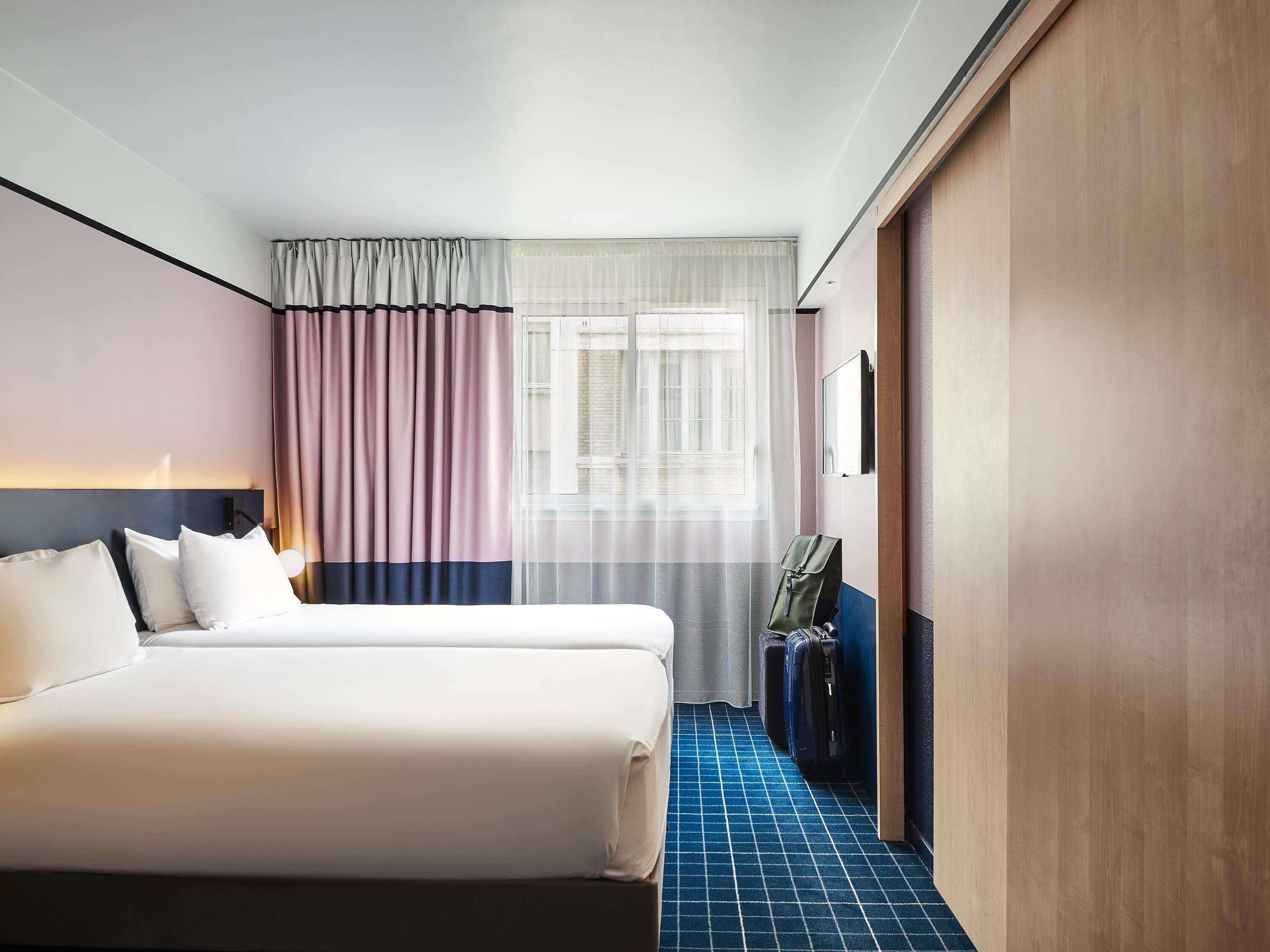 Een hotelkamer met twee bedden, een groot raam met gordijnen, een bureau met stoel en een blauw kamer met een patroon.
