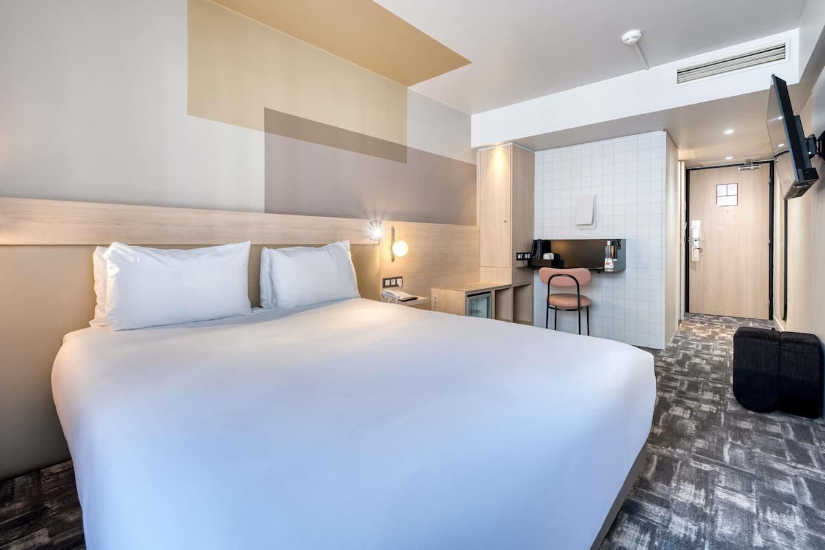 A hotel room with a large bed, a desk with a chair, a small kitchenette, and a TV mounted on the wall.