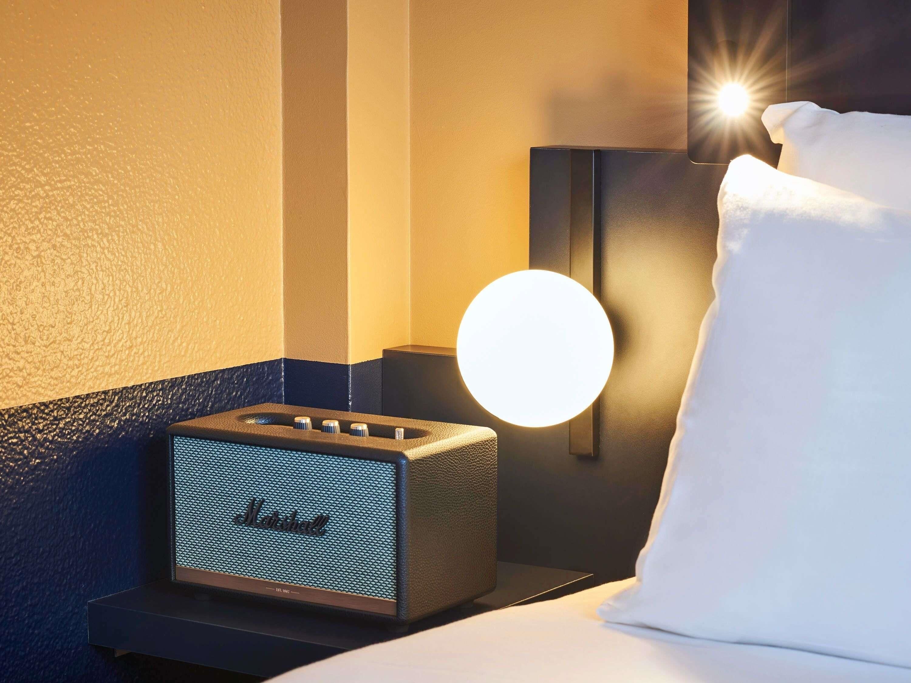 A hotel room with a bed, a nightstand with a lamp, and a wall-mounted speaker.