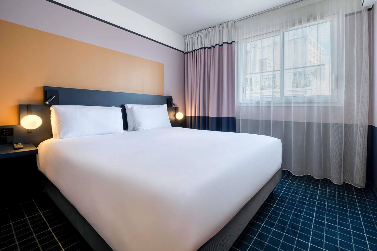 Habitación de hotel con una cama grande, dos mesitas de noche con lámparas, una ventana con cortinas translúcidas y un suelo de patrón azul.