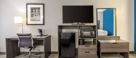 Habitación de hotel con escritorio, silla, cama, televisor, refrigerador y un cuadro en la pared.