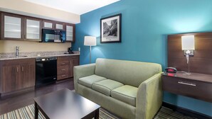 A compact hotel room with a kitchenette, a green sofa, a flat-screen TV, a bedside table, and a nightstand with a lamp. - Best Western Plus Knoxville Cedar Bluff (Knoxville)