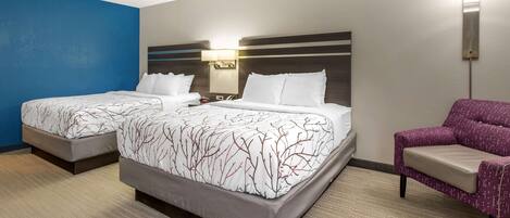 Una habitación de hotel con dos camas, una mesita de noche, una lámpara y un sillón morado.
