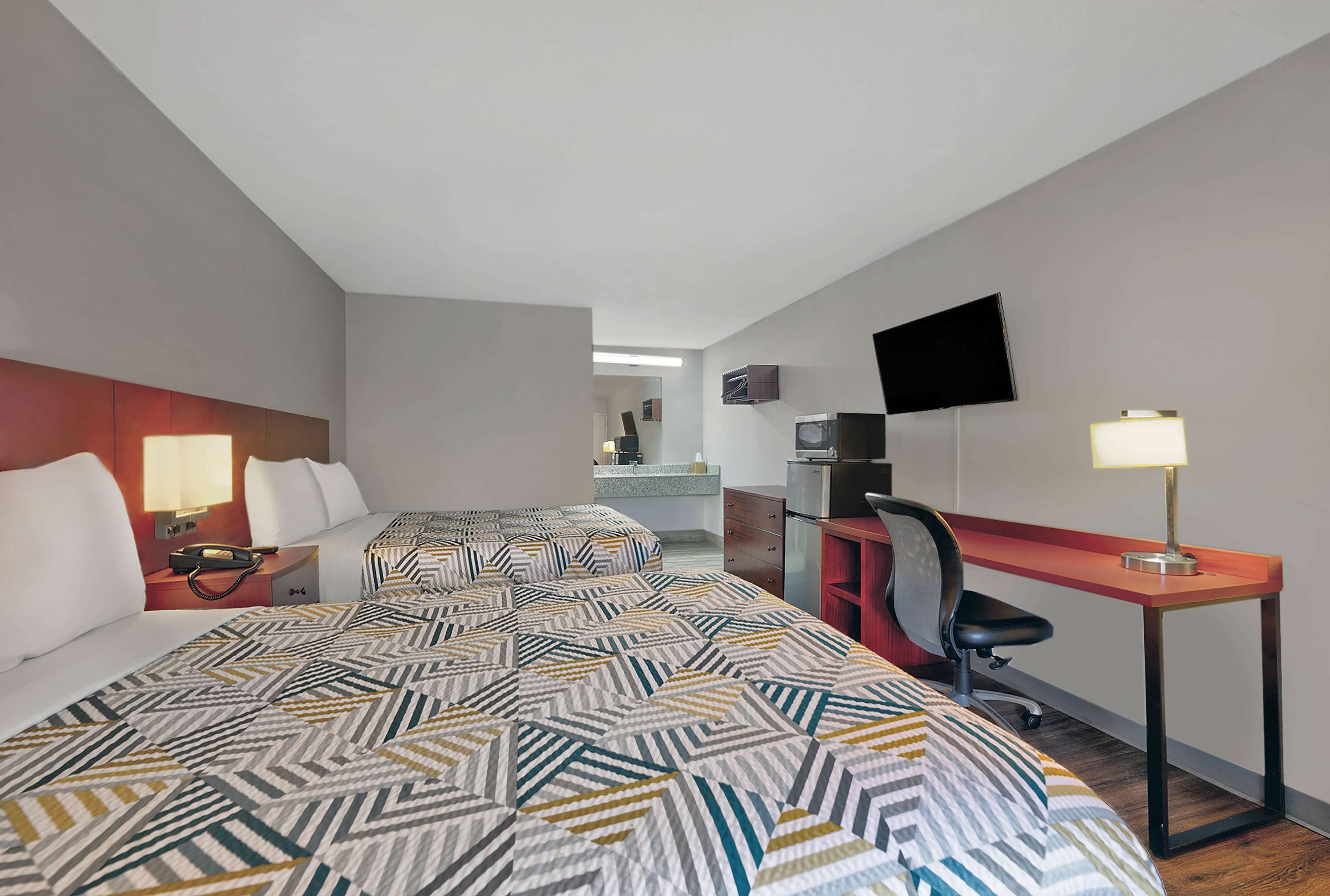 A hotel room with two beds, a desk, a chair, a TV, and a lamp.