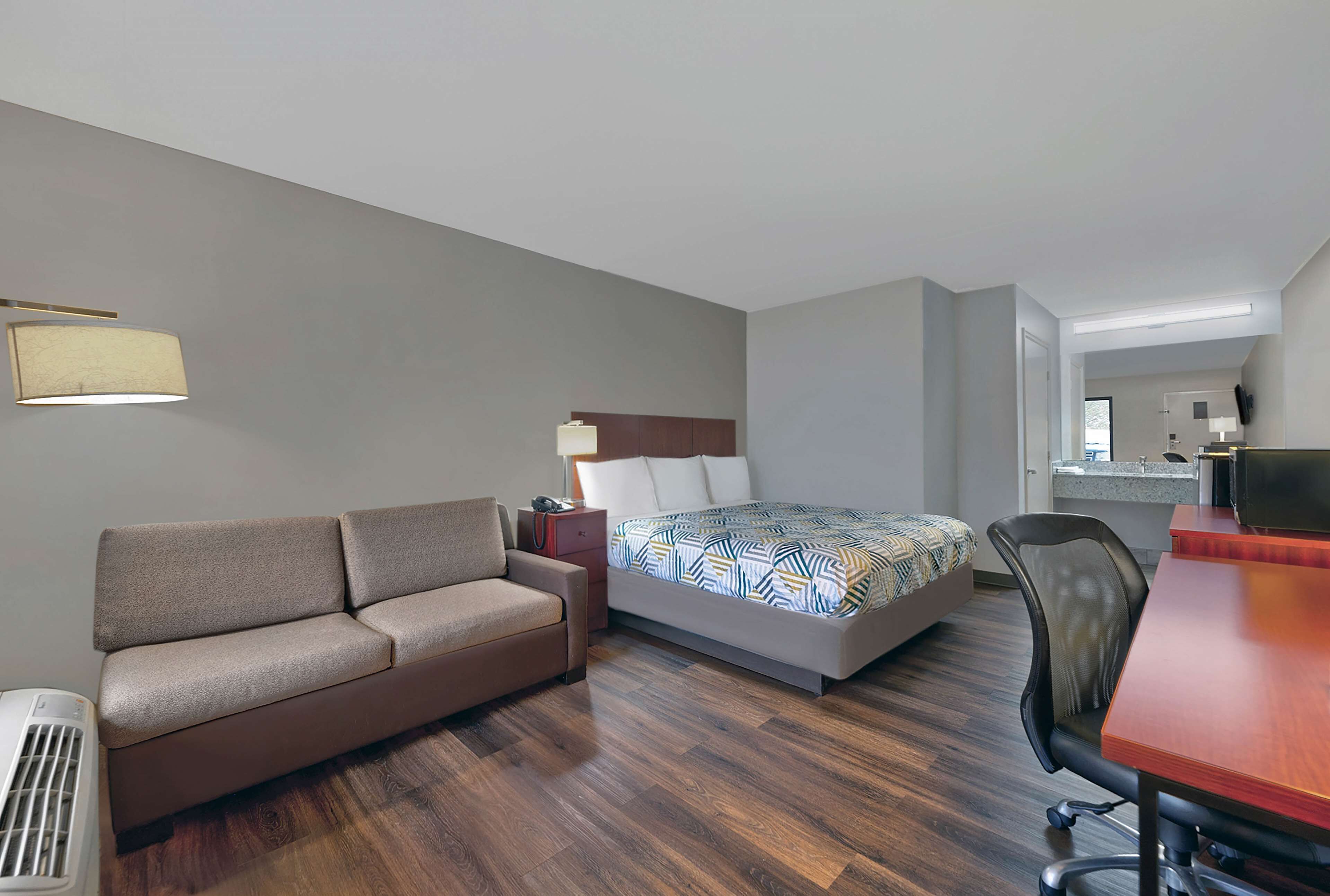 A hotel room with a sofa, a bed with patterned bedding, a desk with a chair, and a kitchenette in the background.
