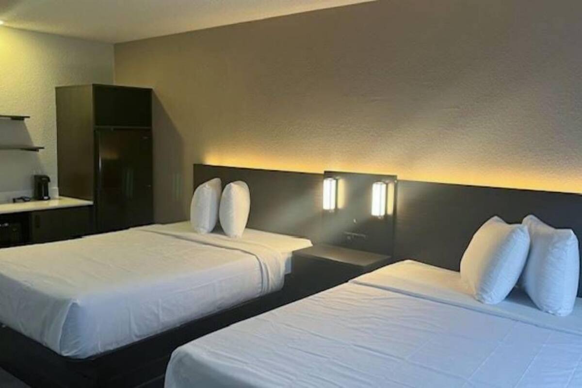A hotel room with two beds, a small desk with a chair, and a kitchenette.