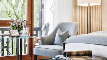 Quarto de hotel com uma cama grande, área de estar com uma poltrona, pequena mesa com um vaso e copos, e janela com vista para o verde.