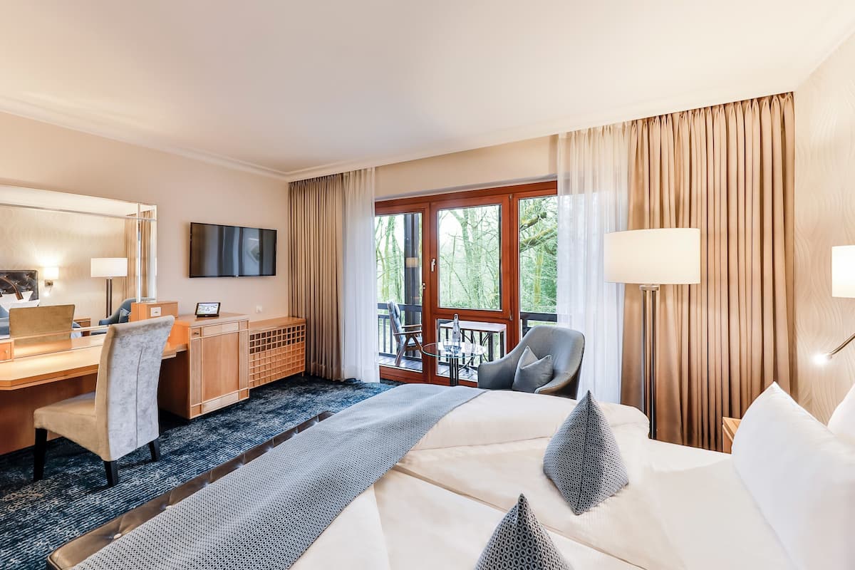 A hotel room with a bed, desk, chair, TV, and a balcony with a view of trees.