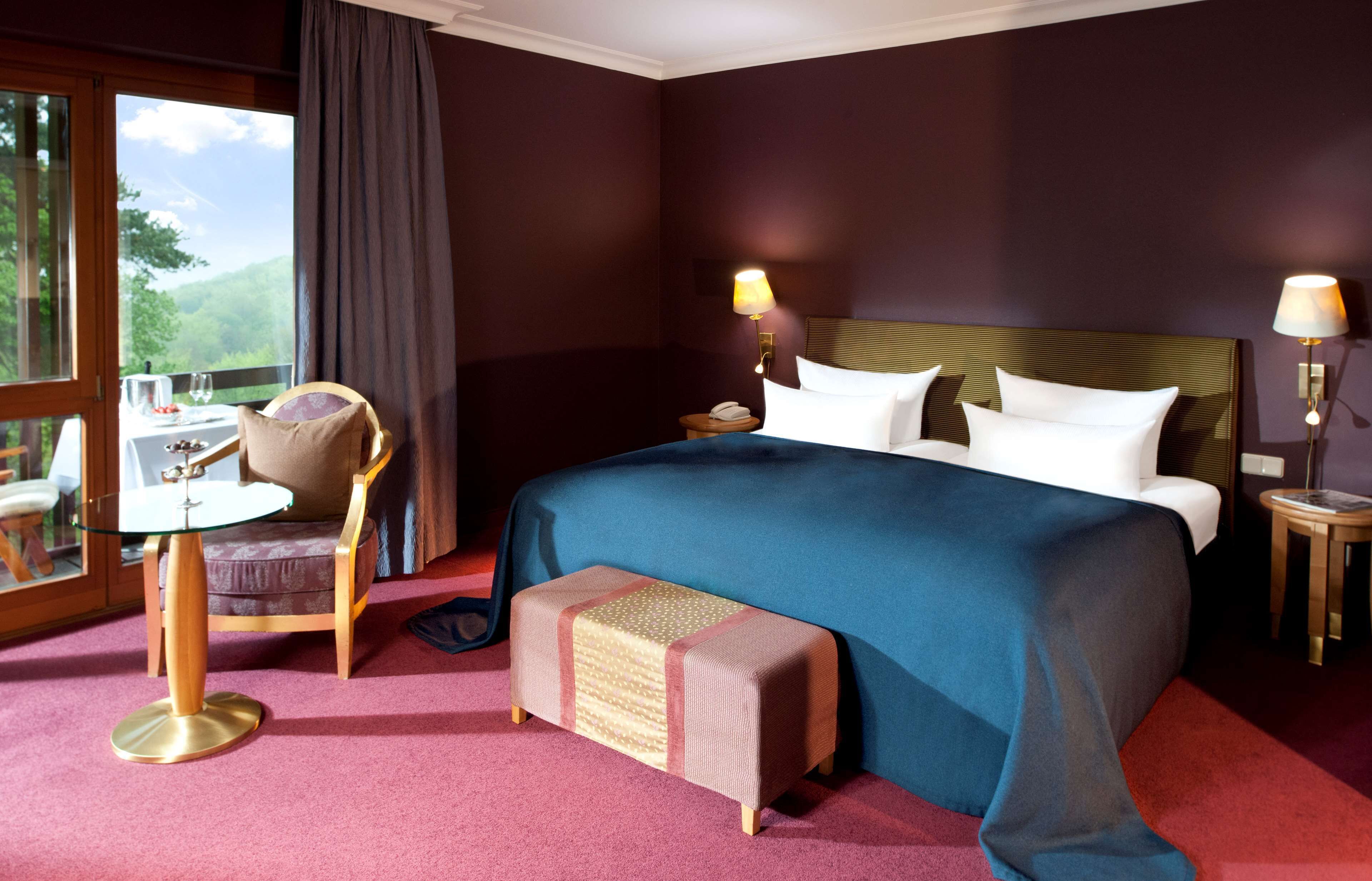 Une chambre d’hôtel avec un grand lit, une petite table ronde, une chaise, un banc et une table de chevet avec des lampes.