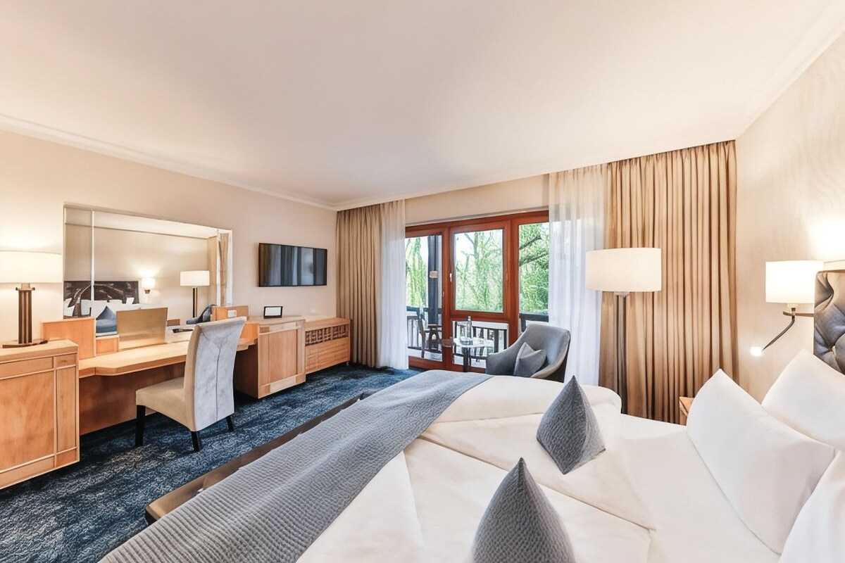 A hotel room with a bed, desk, chair, and a balcony with a view.