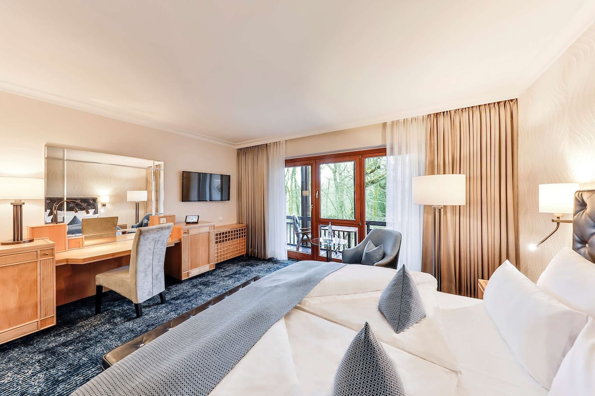 A hotel room with a large bed, a desk with a computer, a chair, a TV, and a balcony with curtains.