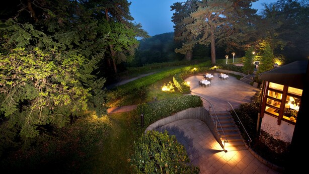 A landscaped outdoor area with a curved pathway, seating arrangements, and a building with illuminated windows. - Dorint Venusberg Bonn (Bonn)