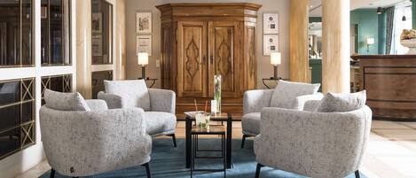 A spacious lobby with two grey armchairs, a small table with a vase and drink, and a wooden cabinet with framed pictures.