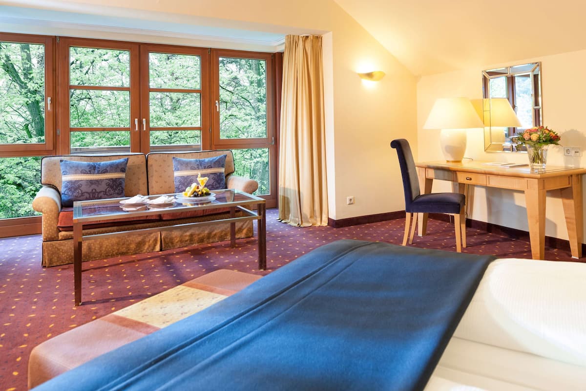 A hotel room with a bed, a desk, a chair, a sofa, and a window with a view of trees.