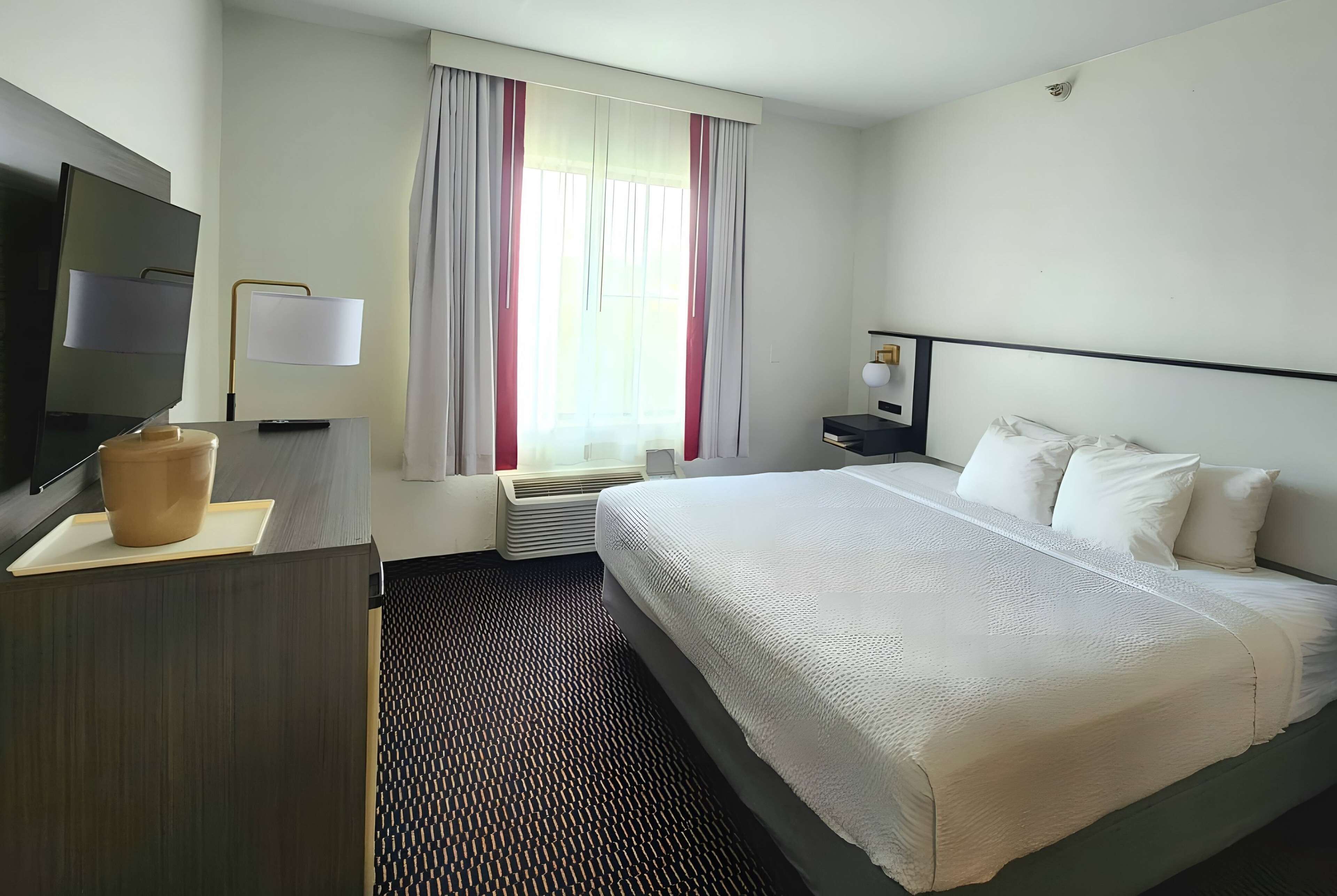 A hotel room with a bed, a desk with a TV, a lamp, and a window with curtains.