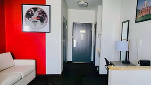A hotel room with a red accent wall, a framed world map, a white sofa, a desk with a lamp, and a door leading to another room. - Ramada by Wyndham Columbia (Columbia)
