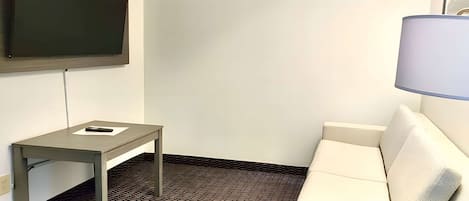 Ein Hotelzimmer mit Flachbildfernseher an der Wand, einem kleinen Tisch mit Fernbedienung, einem weißen Sofa und einer Lampe.