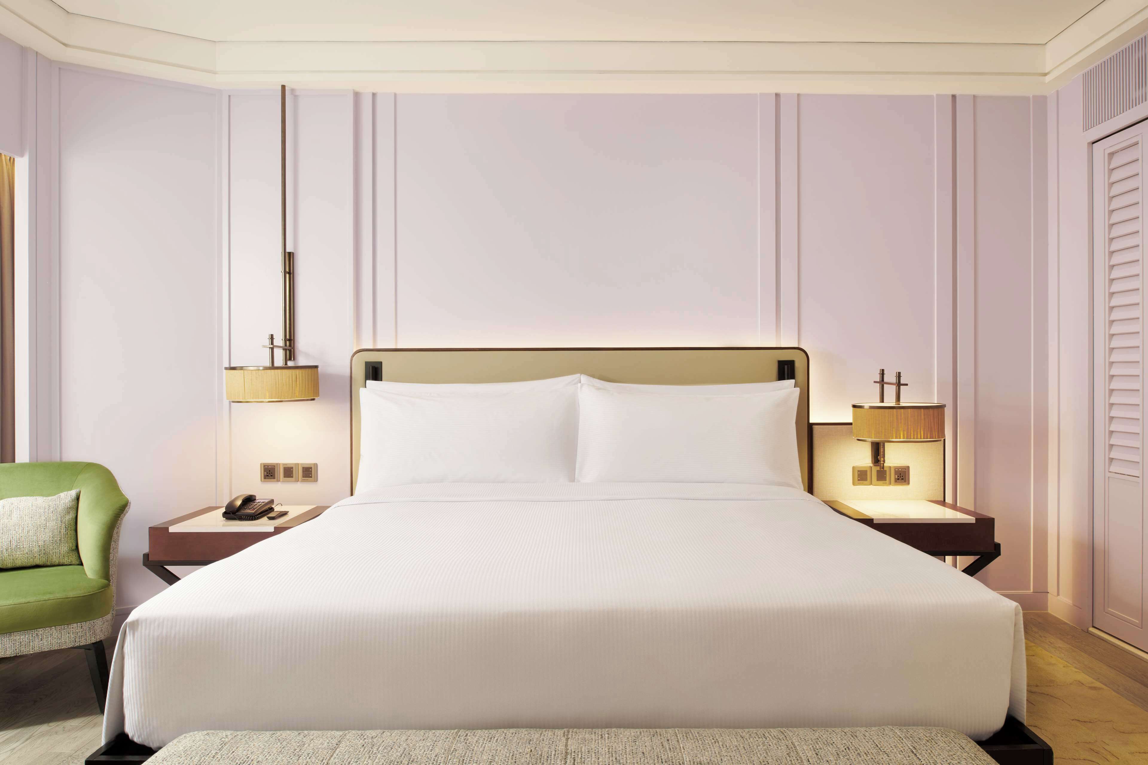 A hotel room with a large bed, two bedside tables with lamps, a green chair, and a wooden floor.