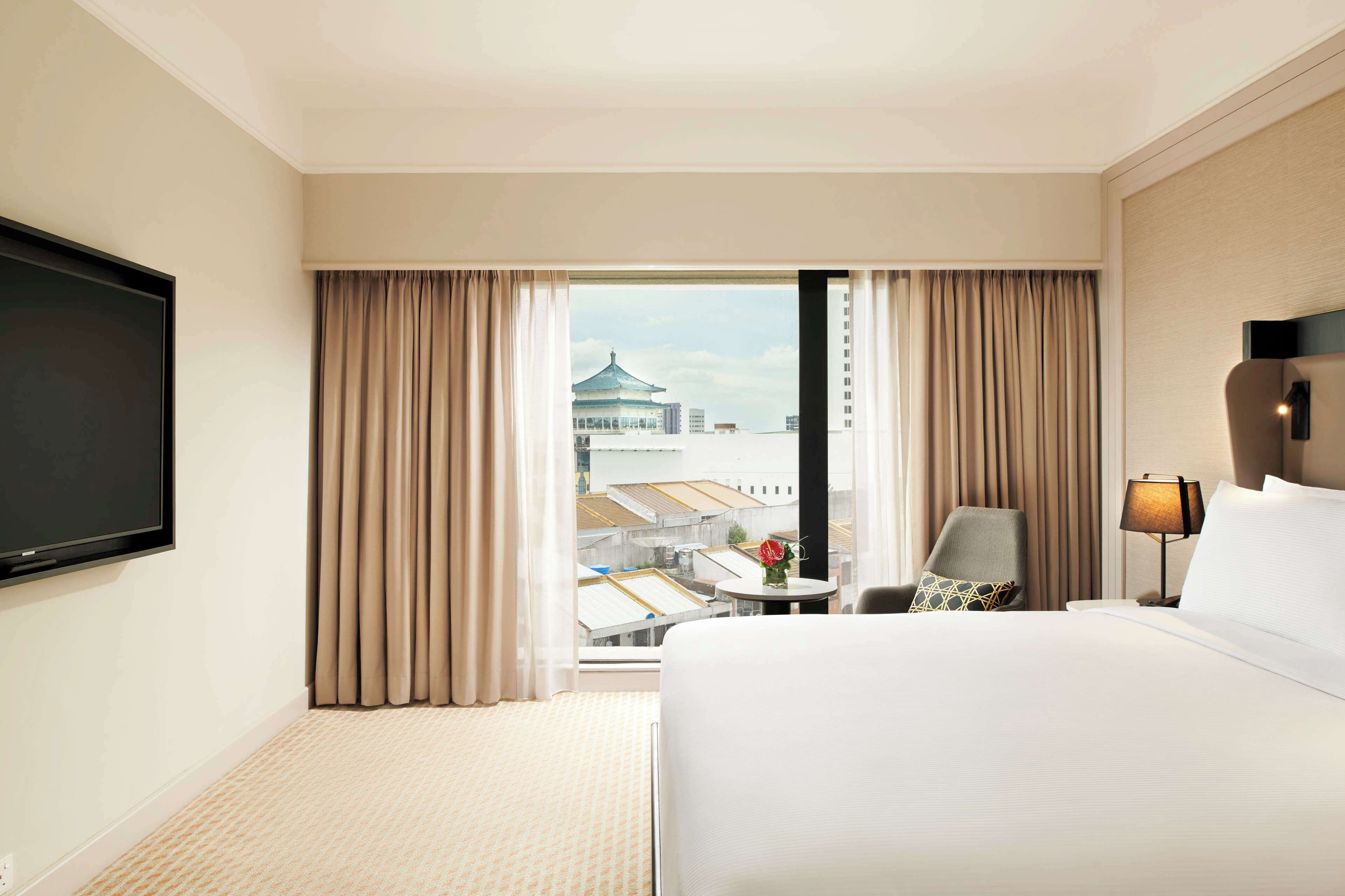 A hotel room with a large bed, a flat-screen TV, a desk with a chair, a lamp, and a view of a building through the window.