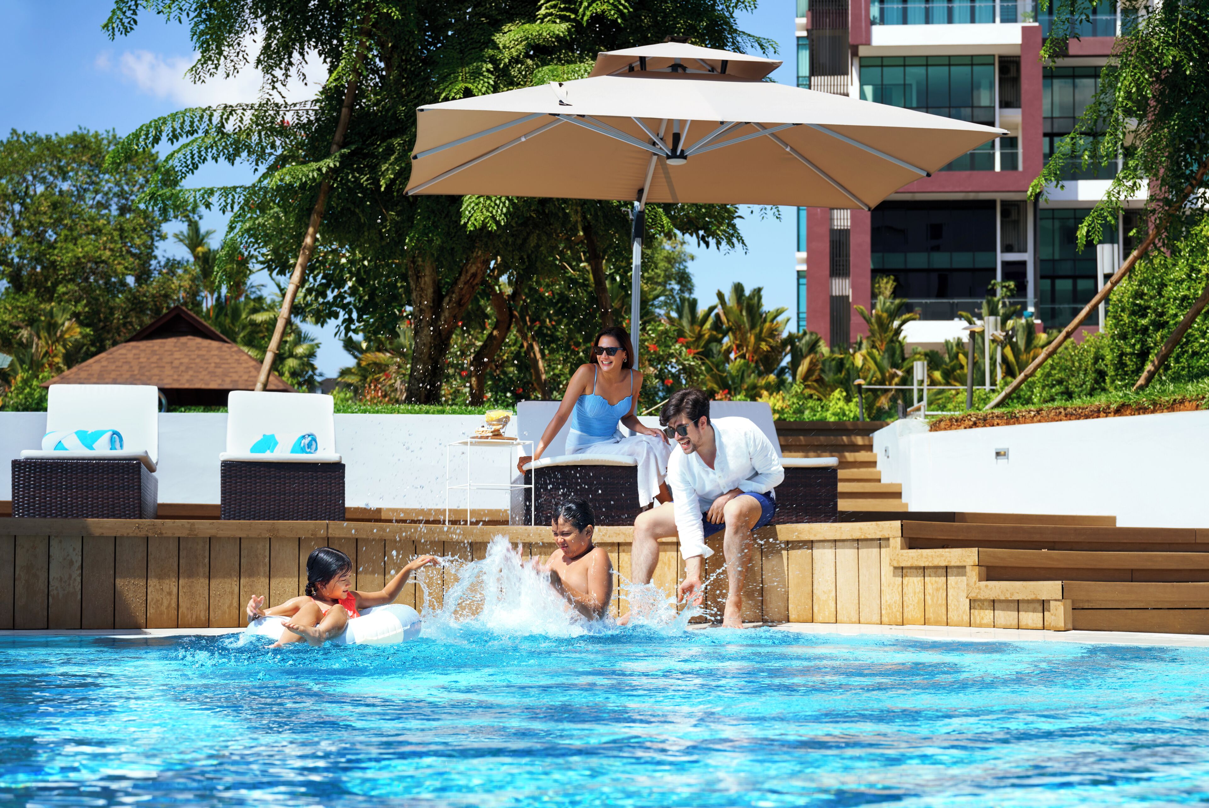 A swimming pool with lounge chairs and an umbrella, surrounded by greenery and a building.