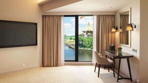 A hotel room with a large TV, a dining table with chairs, a view of a water body and a building with a golden roof, and a large mirror on the wall. - Hilton Kuching (Kuching)