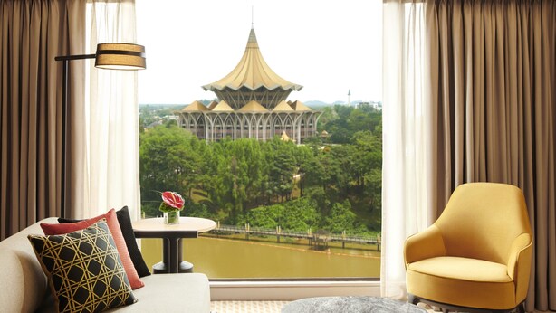 A room with a sofa, a chair, a lamp, and a view of a traditional building through large windows. - Hilton Kuching (Kuching)