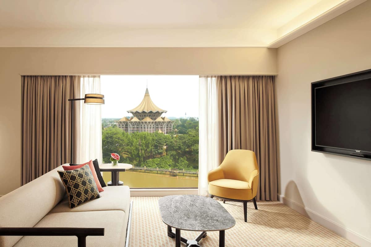 A hotel room with a sofa, armchair, small table, and a TV. A large window offers a view of a pagoda and greenery.