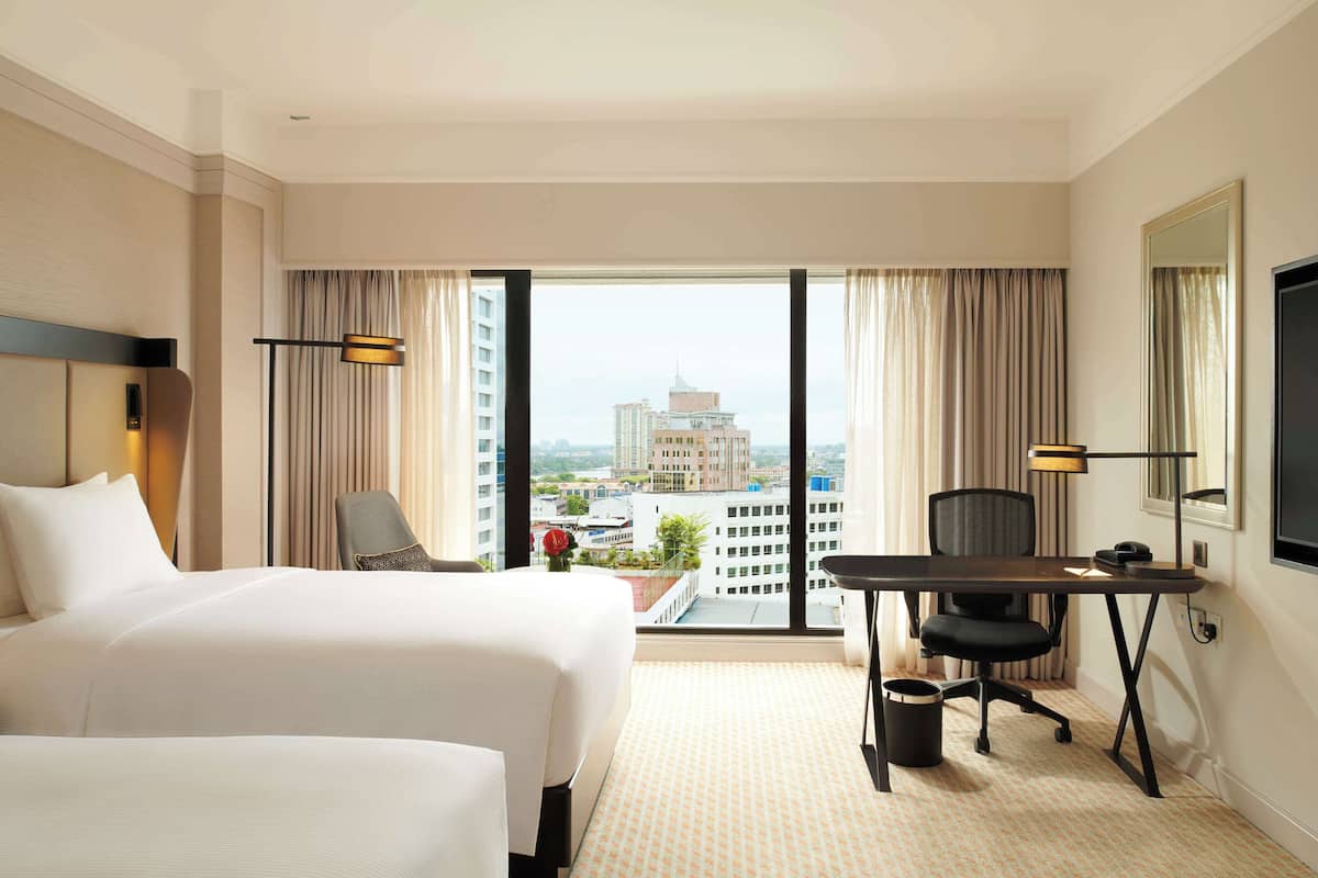 A hotel room with two beds, a desk, a chair, a TV, and a view of the cityscape.