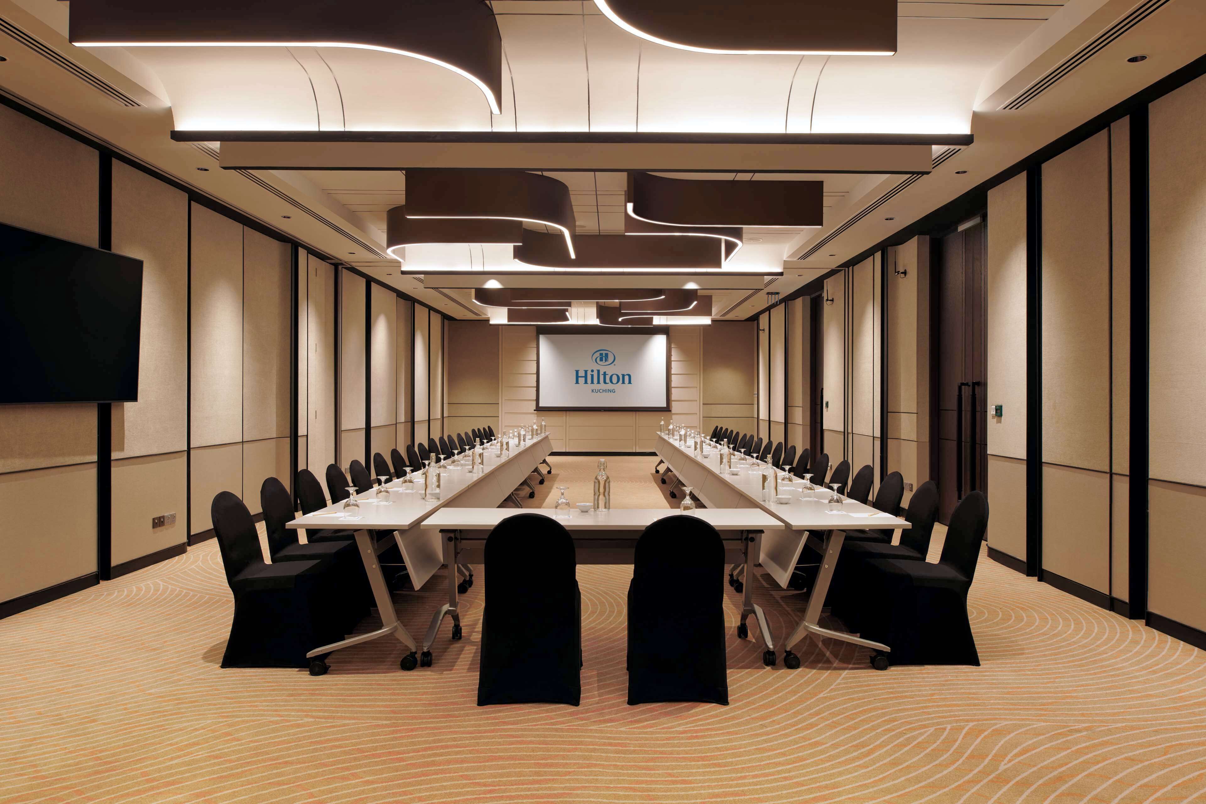 A conference room with a long table, chairs, and a screen displaying 'Hilton Rouching'.