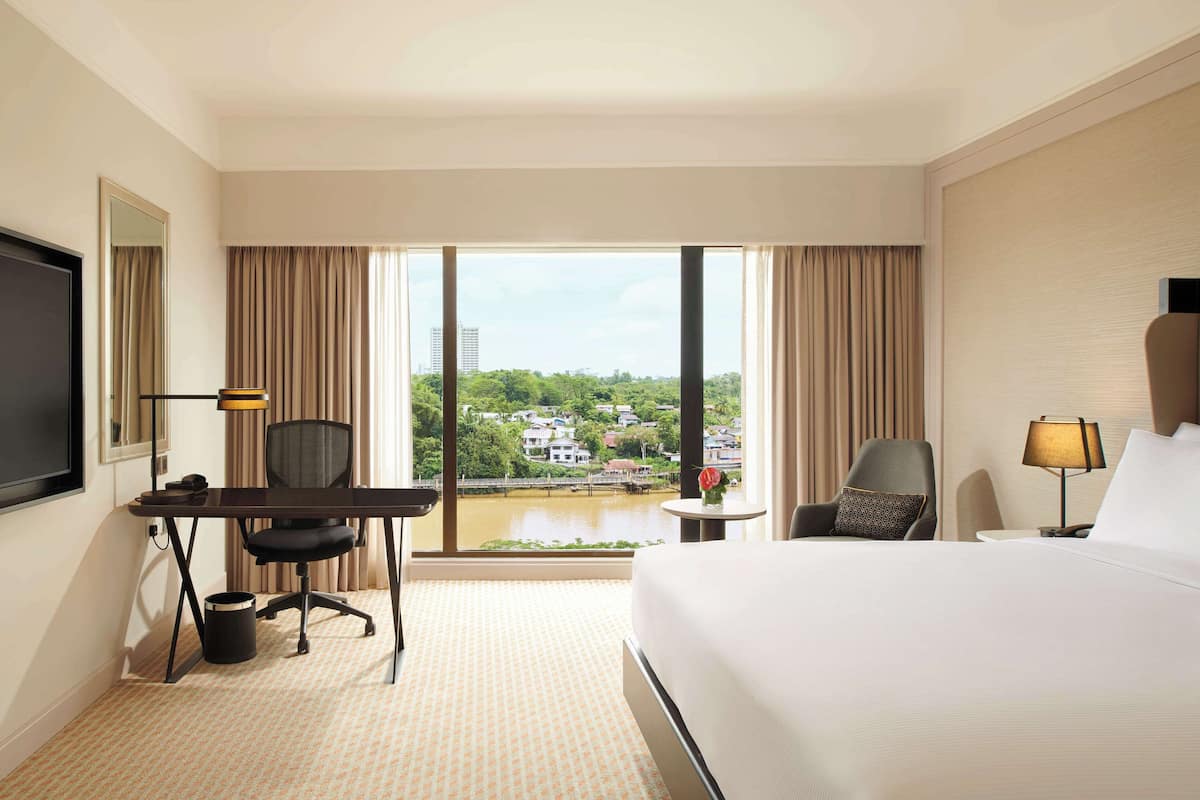 A hotel room with a bed, desk, chair, and a large window with a view of a cityscape and water.