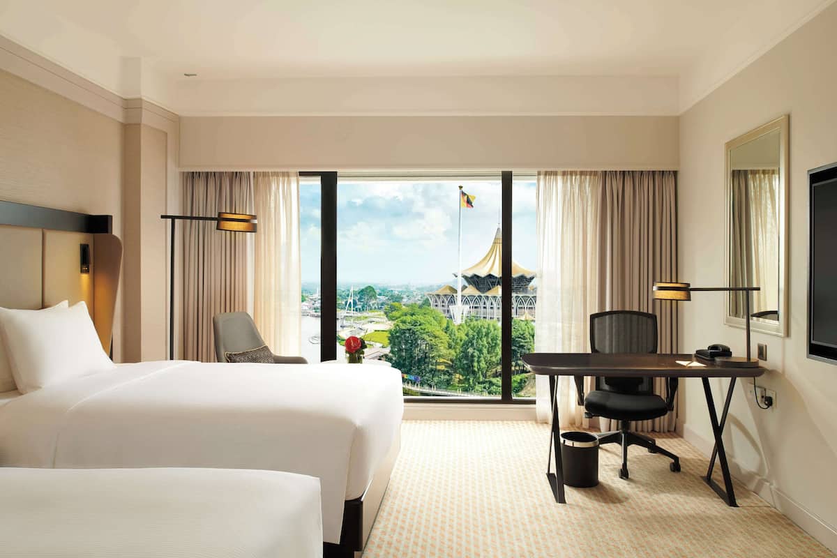 A hotel room with two beds, a desk, a chair, a TV, and a large window with a view of a building.