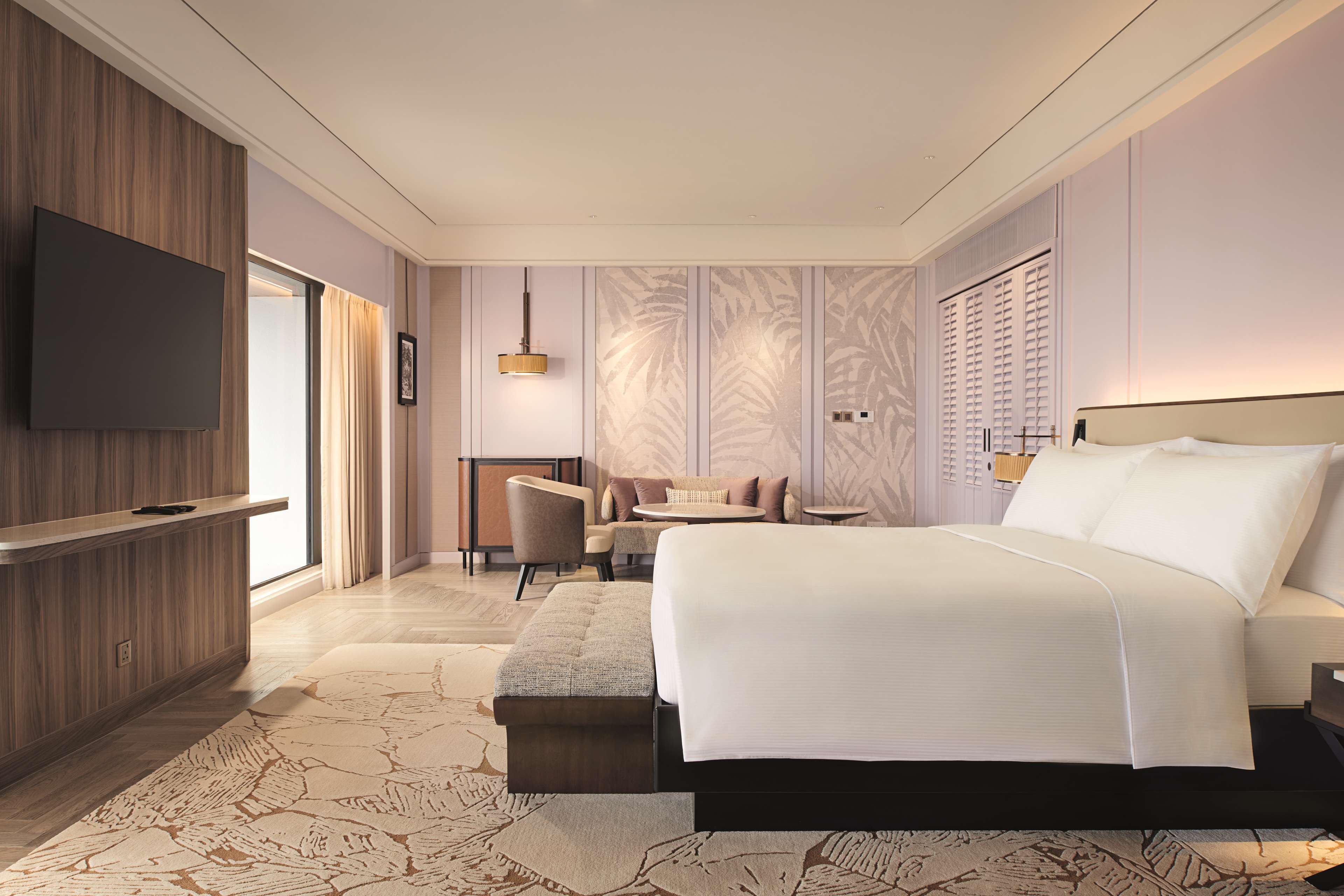 Una habitación de hotel moderna con una cama grande, un televisor de pantalla plana en la pared, un área de descanso con una mesa y sillas, y una alfombra estampada.