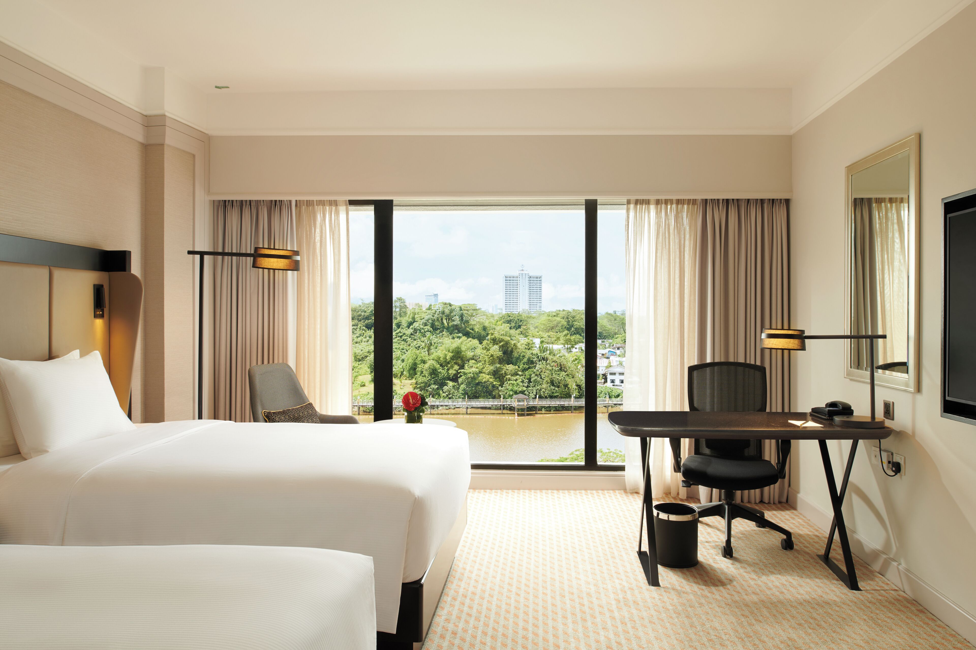 A hotel room with two beds, a desk, a chair, a TV, and a view of a cityscape and greenery.