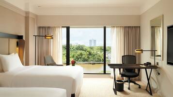 A hotel room with two beds, a desk, a chair, a TV, and a view of a cityscape and greenery.