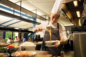 A chef in a professional kitchen with stainless steel appliances, a high ceiling, and a view of diners in the background.