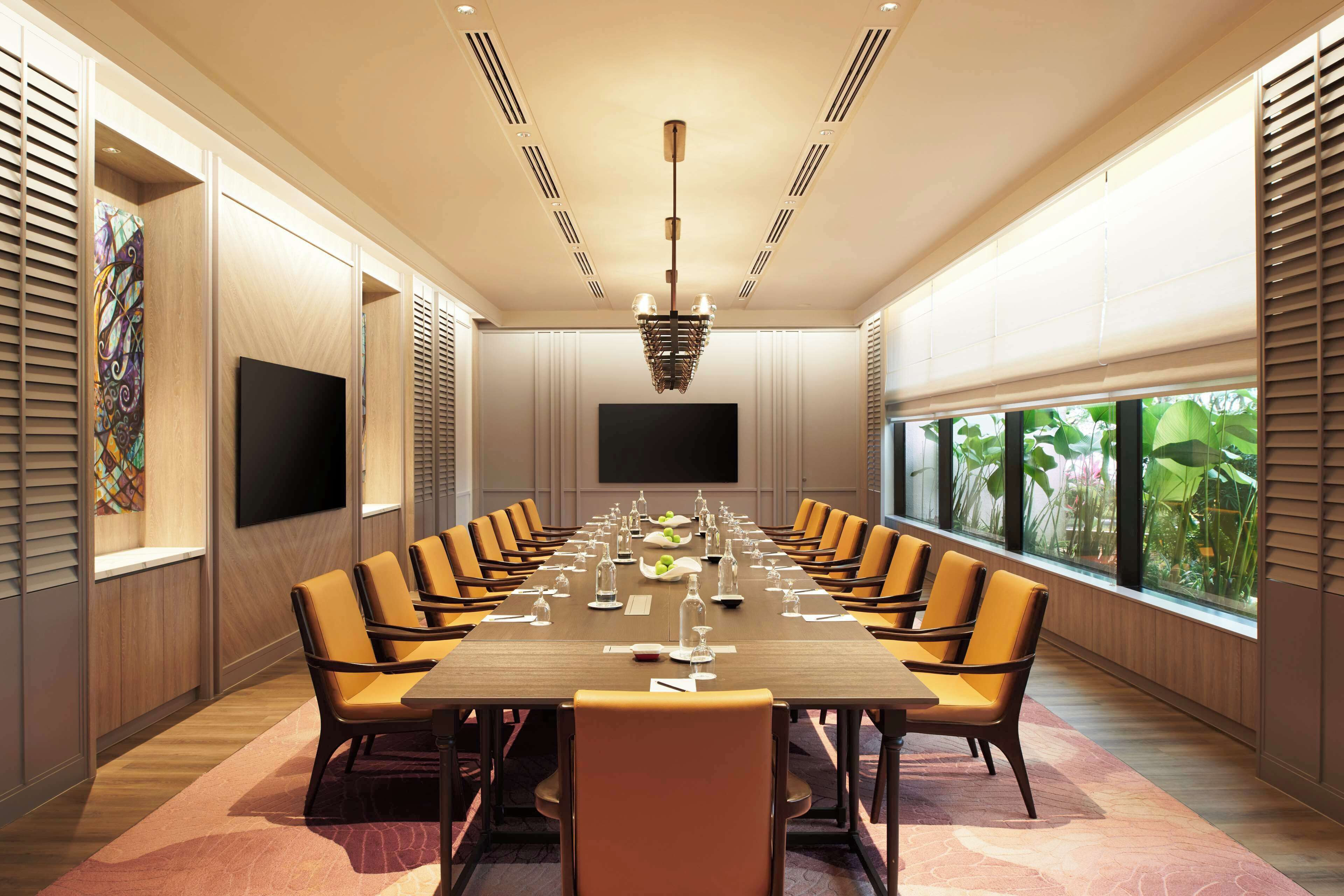 A conference room with a long table, multiple chairs, a flat-screen TV, and large windows with a view of greenery.