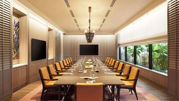 A conference room with a long table, multiple chairs, a flat-screen TV, and large windows with a view of greenery.