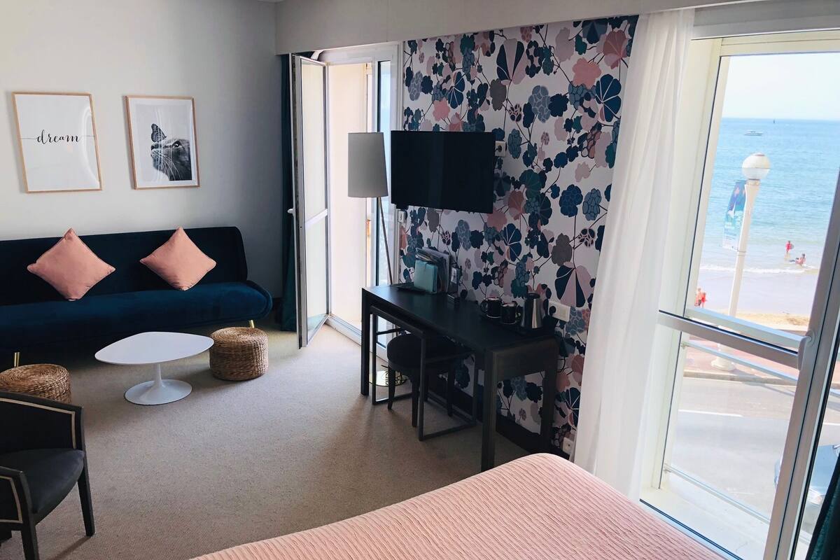 A hotel room with a bed, a desk, a sofa, a TV, and a view of the sea.