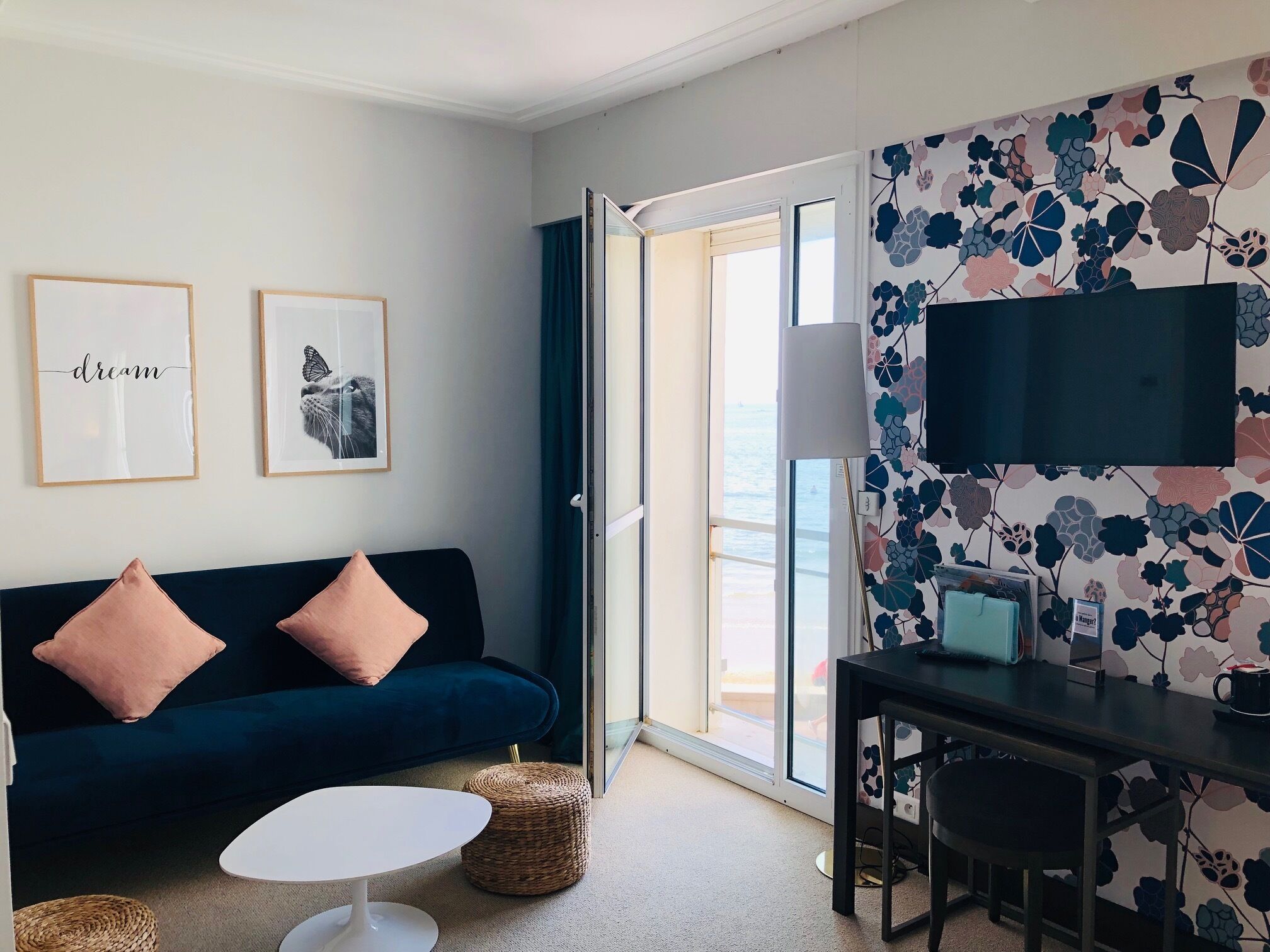 Ein modernes Wohnzimmer mit einem dunkelblauen Sofa, zwei gerahmten Bildern, einem weißen Couchtisch, einem kleinen runden Weidenhocker, einem Schreibtisch mit Stuhl und einem großen Fenster mit Meerblick.
