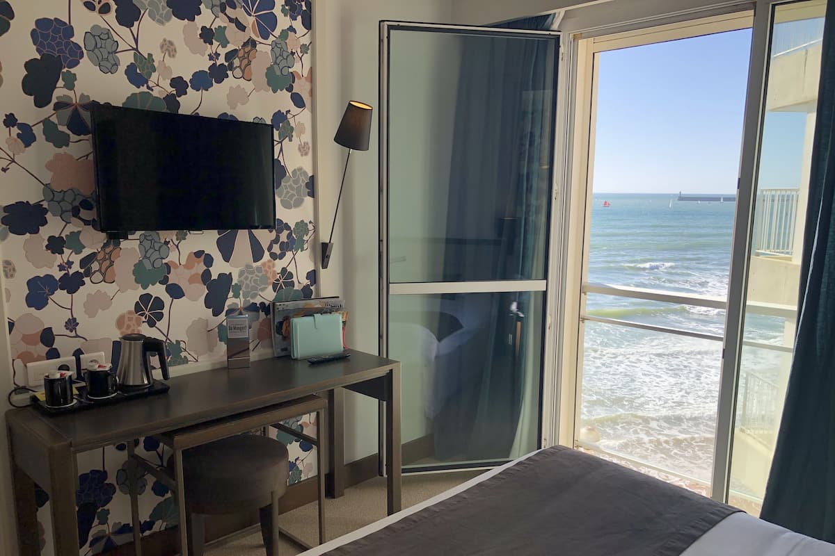 A hotel room with a bed, a desk with a coffee maker, a TV, and a view of the ocean.