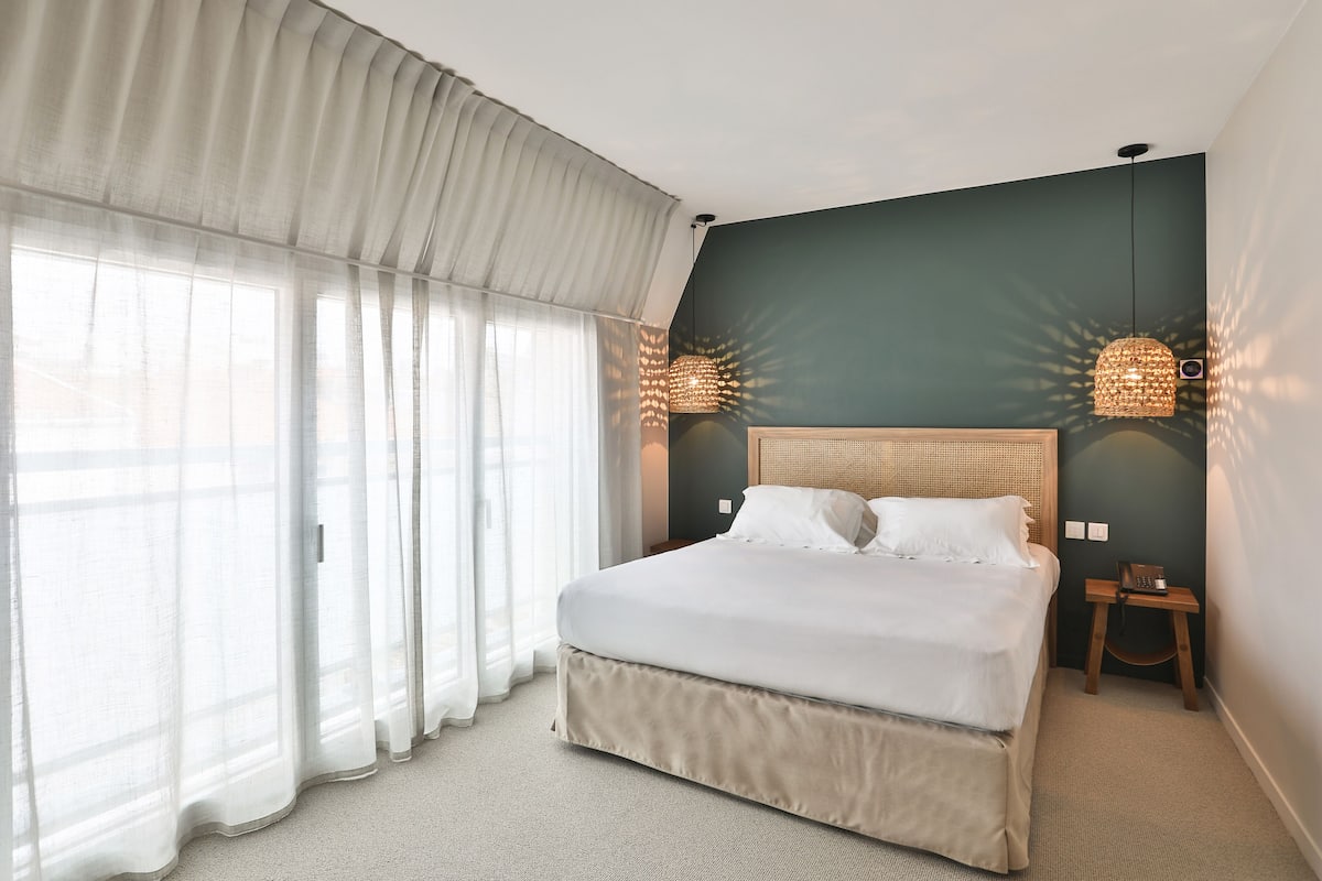 A neatly made bed in a room with a large window, a dark accent wall, and two wall-mounted lamps.