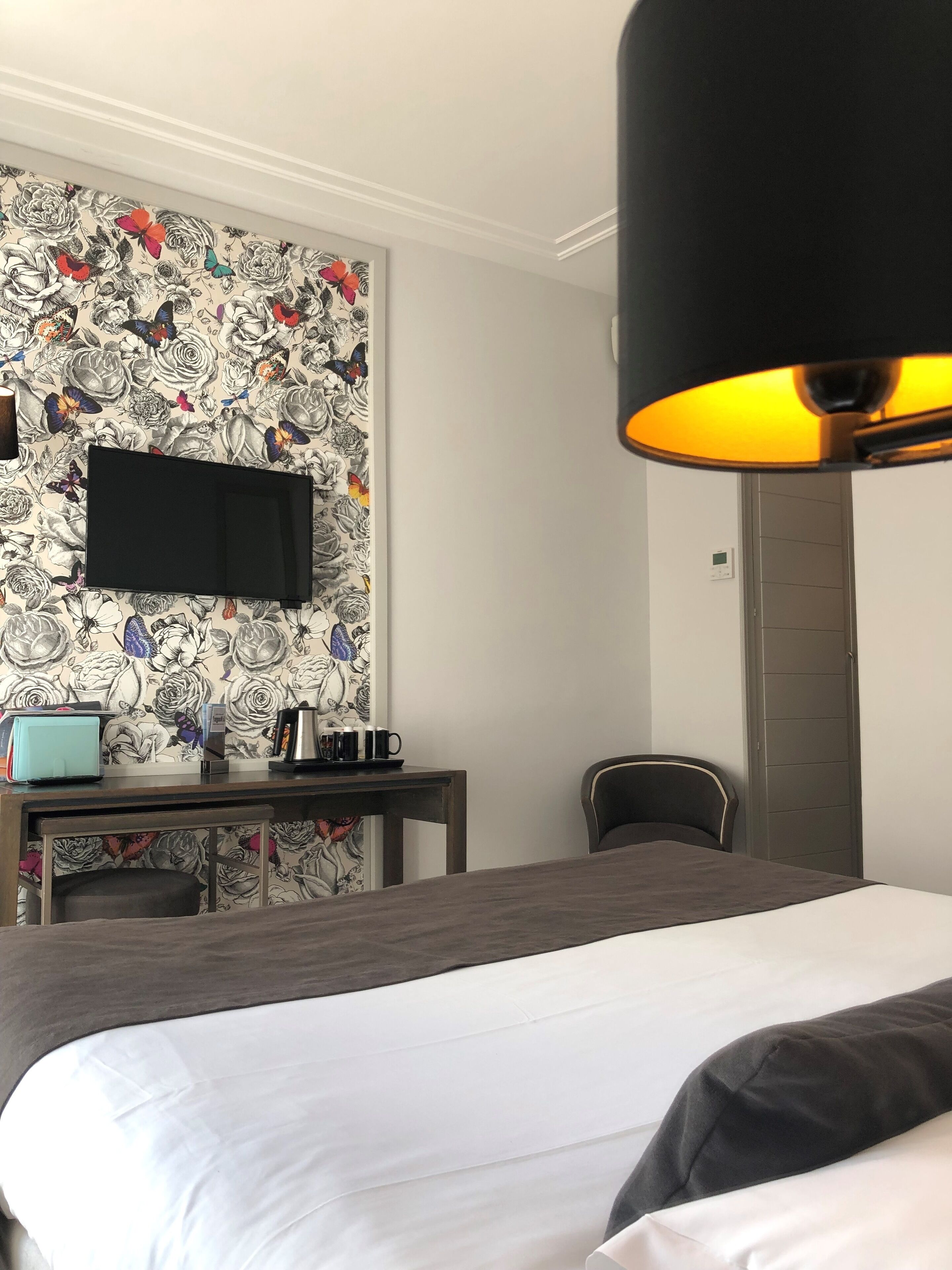 A hotel room with a bed, a desk with a flat-screen TV, a chair, and a wall with a floral pattern.
