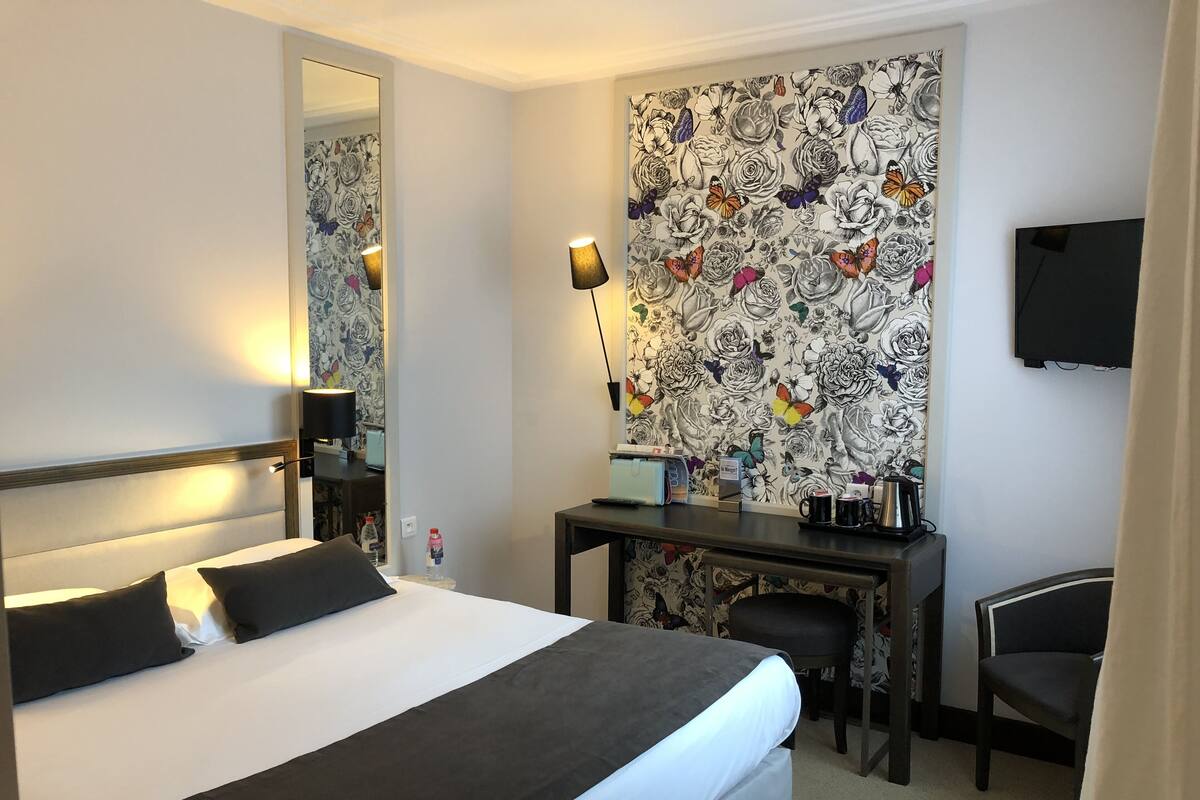 A hotel room with a bed, a desk, a chair, a TV, and a wall with a floral pattern.