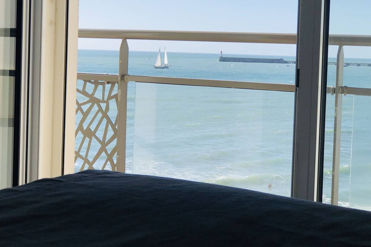 A balcony with a view of the sea, a sailboat, and a distant lighthouse.