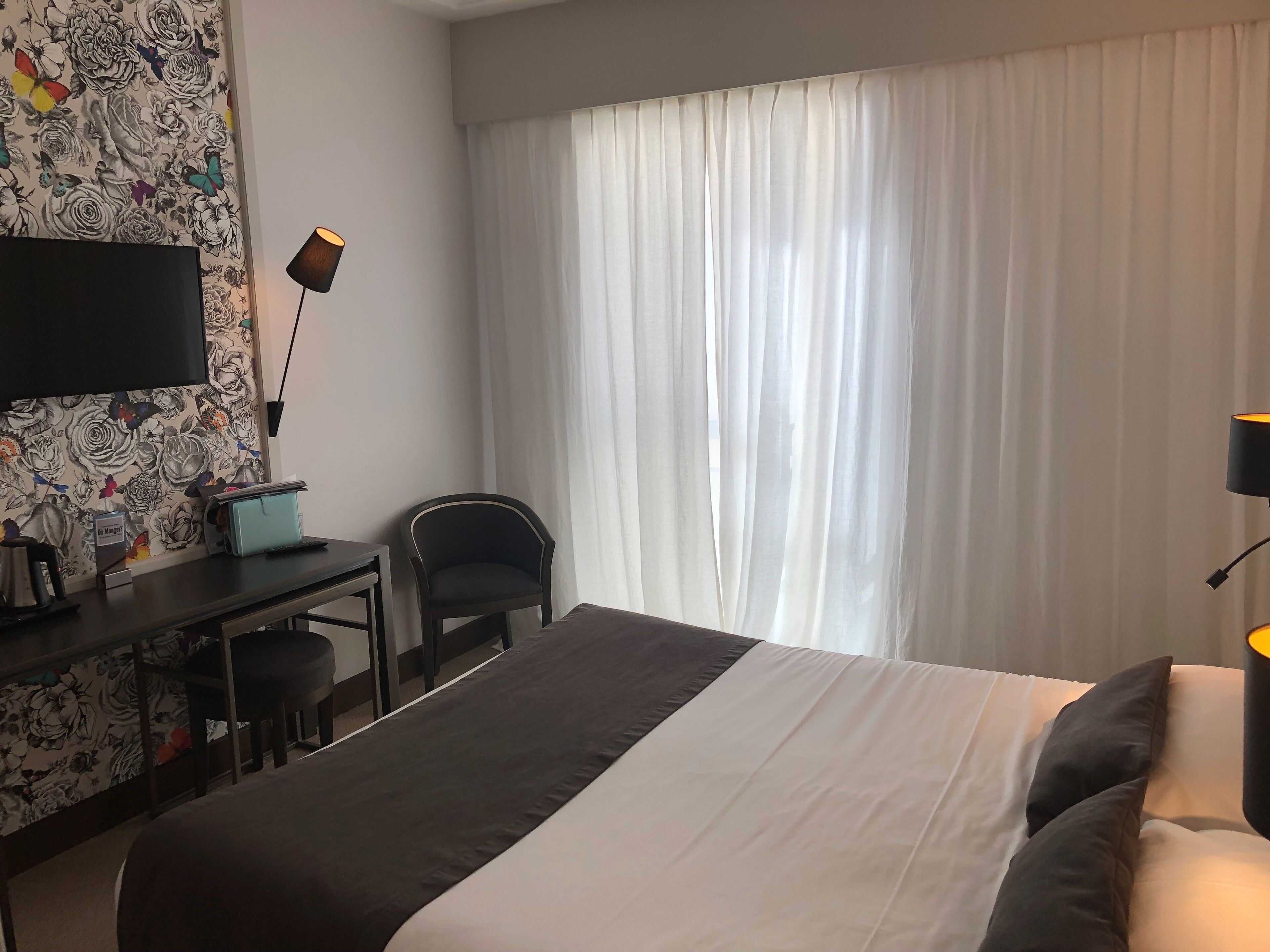 Una habitación de hotel con cama, escritorio, silla, televisor y ventana con cortinas.