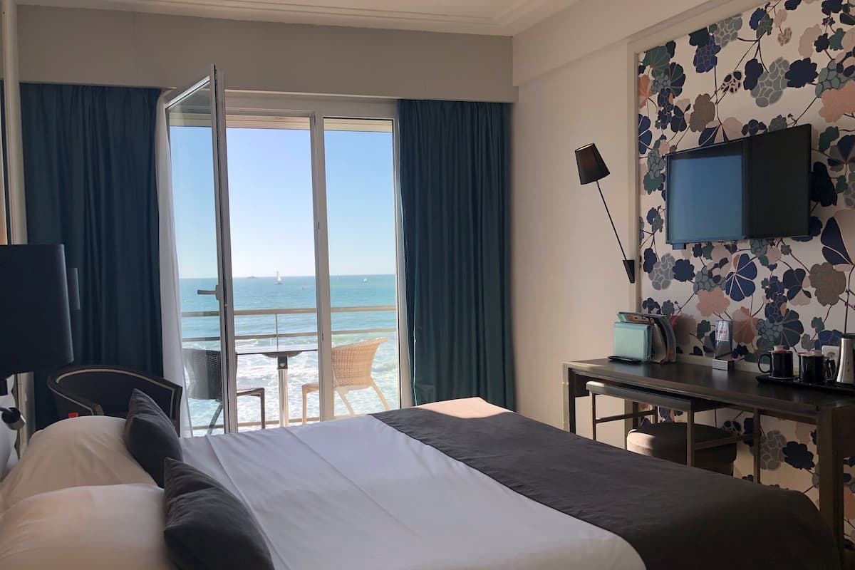 A hotel room with a bed, a desk, a chair, a TV, and a view of the sea.